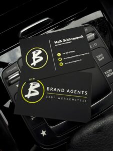 Brand Agents Visitenkarten mit gelbem Logo auf schwarzem Hintergrund, Maik Schönepauck genanntes Branding, kompakte Karten im Autoinnenraum.